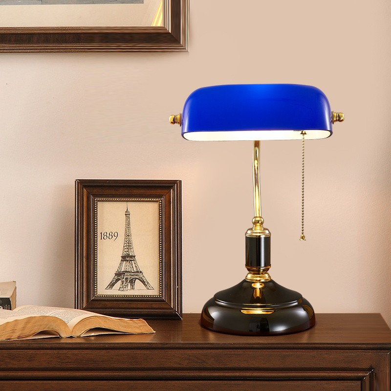 Blue banker lamp on wooden desk with books and framed artwork