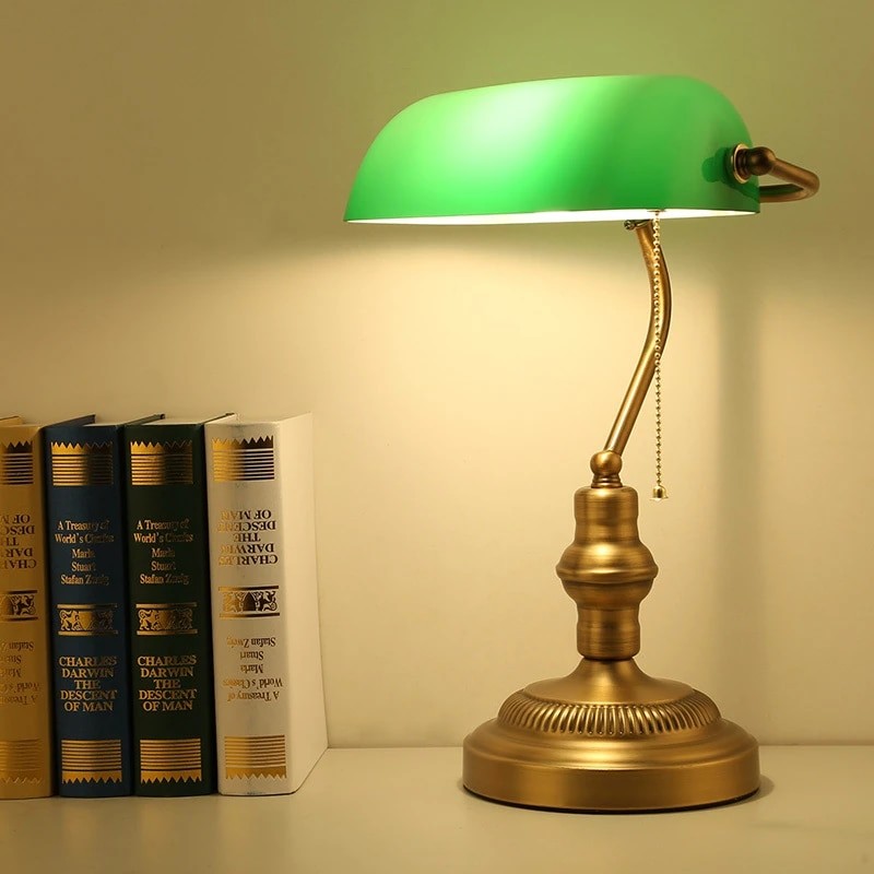 Green banker lamp lit beside books with brass base